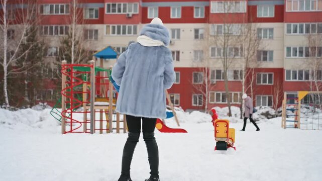 Mom and son playing in snow at urban playground courtyard, mother lifts and spins child while bundled in coat hat and scarf, joyful smiles and laughter, apartment buildings in background, closeup