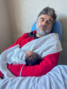 Man Sleeping In Hospital Chair Holding Newborn Baby
