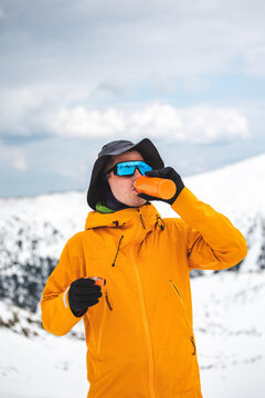 Man in an orange jacket putting on skis, drinking water 