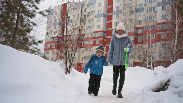 Mother son walking along snow path near apartment blocks, pine trees and tall buildings framing scene, child in blue jacket and hat, mother carrying green sled and wearing warm gray coat with white