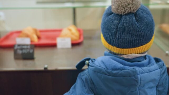 Little white boy looks up questioning, asking mom for pastry behind glass case, blue jacket and pompom hat, bakery counter scene, hopeful expression and gentle conversation, waiting permission