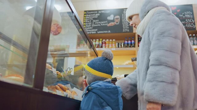 Mom and son at bakery counter choosing pastries bundled in winter coats and knit hats mother points at croissants behind glass display warm indoor lighting chalkboard menu and shelves in background