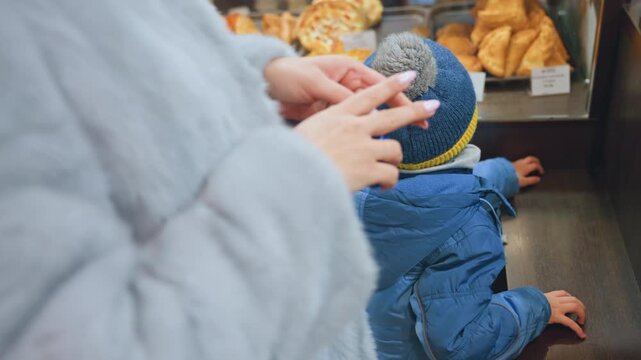 Mom and son paying at bakery, child watches mother tap card at counter, closeup of hands and payment card, pastries in display case, toddler in blue coat and pompom beanie leans on counter, candid
