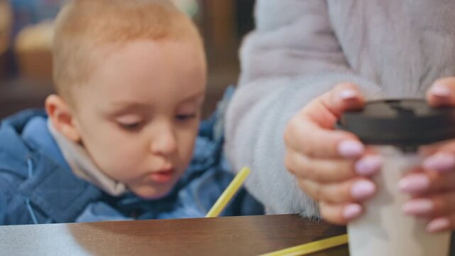 White mom and son share coffee moment, toddler reaches for lid while mother steadies cup with manicured nails on cafe table, cozy warm lighting, protective parental gesture and quiet morning routine
