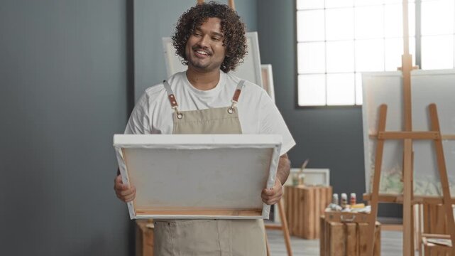 Man holding blank canvas with both hands, smiling and cradling it, wearing apron, in art studio with easels, paint bottles and wooden stools by window; creative joy.