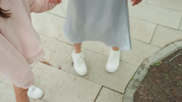 White mom and daughter pause closeup shoes on pavement, matching white sneakers and pink hoodie, gentle handhold and small talk, soft pastel tones and urban curbside framing, intimate family moment