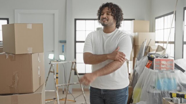 Man with arms crossed beside stacked moving boxes in building, casual white tshirt and jeans; home settling contentment.
