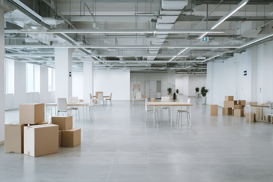 Large, bright, and mostly empty interior space with concrete floor. Cardboard boxes, tables and chairs are scattered throughout the area