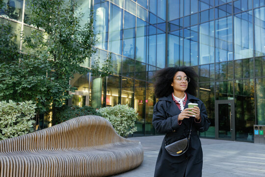 Black businesswoman with coffee going near glass skyscraper building