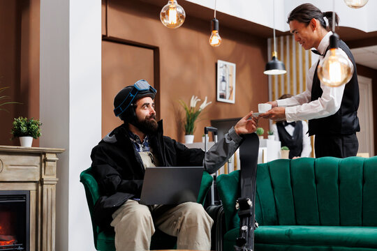 In hotel lobby asian waiter attentively brings refreshing beverage to tourist lounging on couch with digital pc. Male guest wearing winter jacket receiving cup of coffee from employee in ski resort.