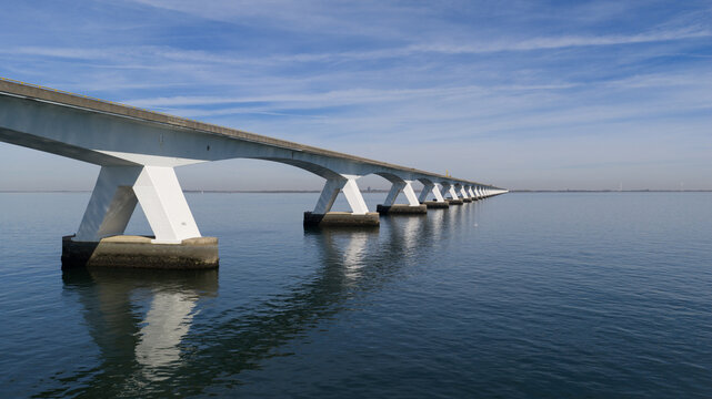 Aerial view of the Zeeland Bridge with its long concrete structure and white V-shaped pillars stretching over calm blue water under a clear sky in Zierikzee, Zeeland, Netherlands.