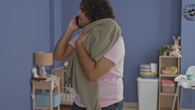 Man cradles a bundled blanket to his chest and talks on smartphone amid a crib and nursery toys in a baby room building; parental concern.