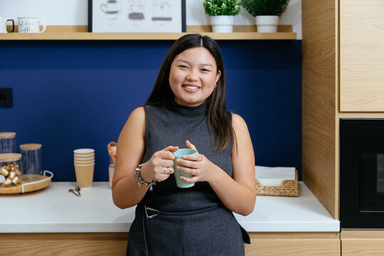 Young asian woman smiling holding mug in office kitchen