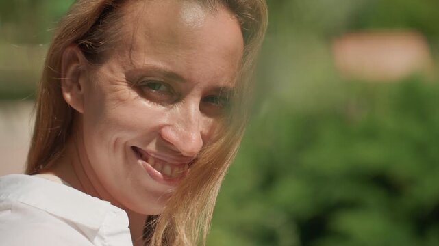 White mom smiles warmly at camera with daughter beside, sunlight on face, relaxed confident gaze, natural beauty, garden background, candid maternal portrait, lifestyle warmth, authenticity