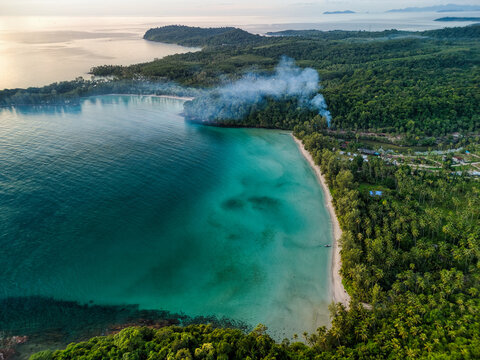 Aerial view of the turquoise waters and white sand beach of Ko Kut with smoke rising from the dense tropical forest in Ko Kut District, Trat, Thailand.