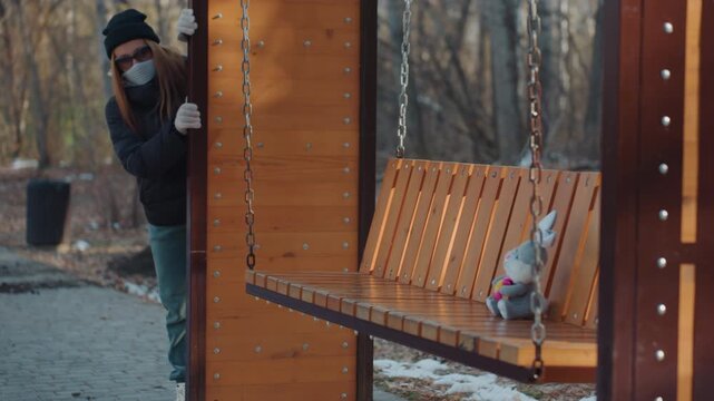 Caucasian woman snatching teddy on bench, park swing seat with chain and wooden slats, backpack over shoulders, gloved hands grab stuffed toy then lift toward pocket, furtive glance and hurried motion