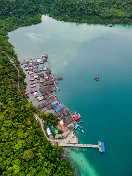 Aerial view of a traditional fishing village with wooden stilt houses along the turquoise coastline and lush tropical forest in Ko Kut District, Trat, Thailand.
