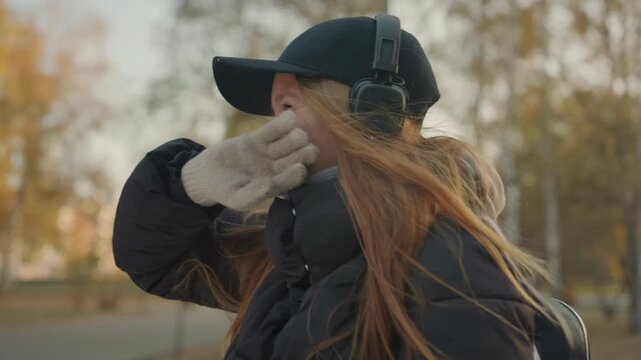 Smiling caucasian woman with headphones enjoying autumn park breeze, wearing black cap, puffy jacket and knit gloves windswept red hair and upbeat laughter create warm candid mood frame shifts suggest