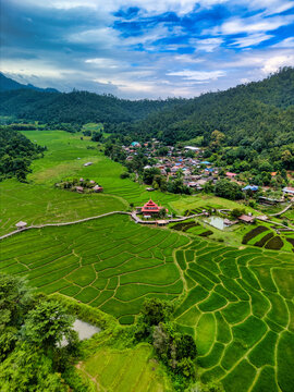 Aerial view of Kho Ku So Bamboo Bridge stretching across lush green rice terraces and a small village surrounded by forested hills in Thung Yao, Mae Hong Son, Thailand.