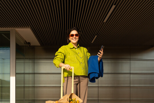 Woman stands with suitcase and phone at modern building entrance