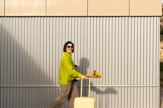 Woman walks with luggage against a colorful wall during the day