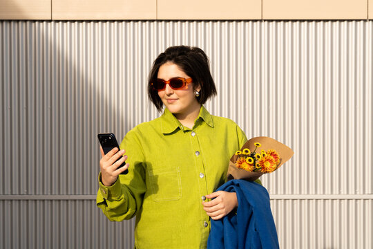 Woman holding flowers and phone while standing in front of wall