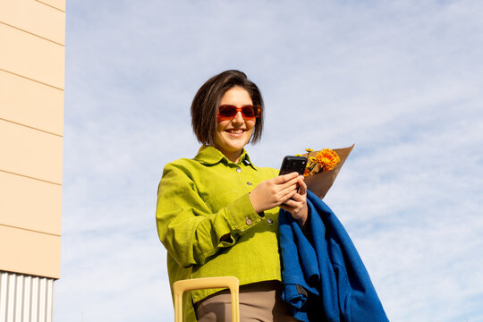 Woman smiles while holding a phone, flowers, and coat outdoors