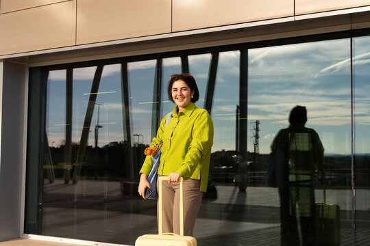 Woman with suitcase smiles near large windows in vibrant setting