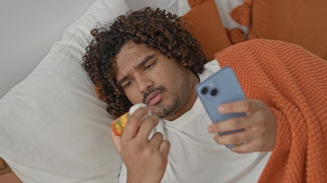 Man in bed holding smartphone and prescription pill bottle, finger tracing the label while checking phone for dosage info in a building; concern.
