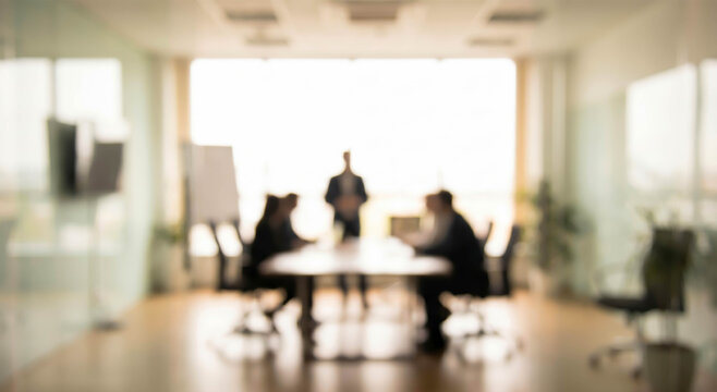 A blurred image of a business meeting in a modern office space, showing individuals engaged around a conference table with a presenter standing.