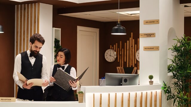 Happy receptionists cross referencing tourists booking appointment data at hotel reception desk. Teamworking professional resort staff working at check in counter in stylish holiday destination