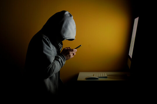 Anonymous hacker using smartphone while working on computer in dark environment, representing mobile hacking, cybercrime and data theft.