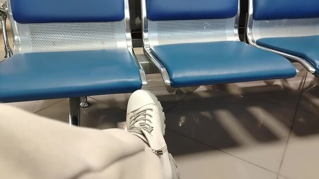 Foot of a person in white sneakers resting on a blue airport chair, showcasing a relaxed posture in a modern terminal setting with additional empty seats visible