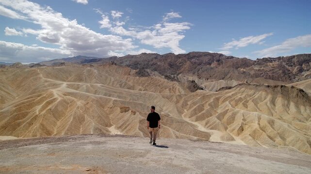 A man in black shirt stands at Zabriskie Point viewpoint gazing over dramatic eroded badlands and colorful sedimentary rock formations under blue sky with clouds. Death Valley National Park.