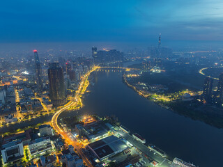 Obraz premium Aerial evening view over Saigon River with glowing roads, dense high-rise buildings, and atmospheric haze covering Ho Chi Minh City skyline.