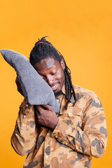 Fototapeta premium Dreamful person holding pillow to take a nap in studio, comfortable pose to sleep and rest. African american young adult with eyes closed trying to fix slpeeing schedule with bedtime hour.