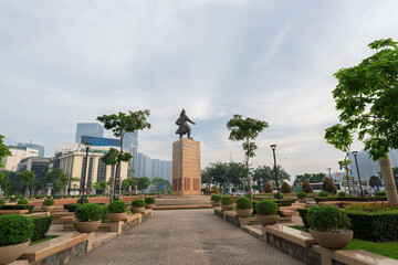 Obraz premium Tran Hung Dao statue monument centered in landscaped park with symmetrical pathways, trimmed shrubs, and modern skyline backdrop under cloudy sky in Ho Chi Minh City.
