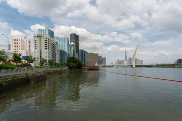 Obraz premium Riverside cityscape with modern buildings and distinctive bridge structure, calm water surface, and cloudy sky in developing urban district.