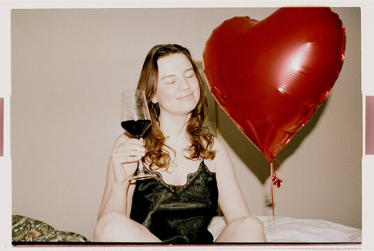 Smiling Brunette With Red Wine and Heart Balloon on Bed