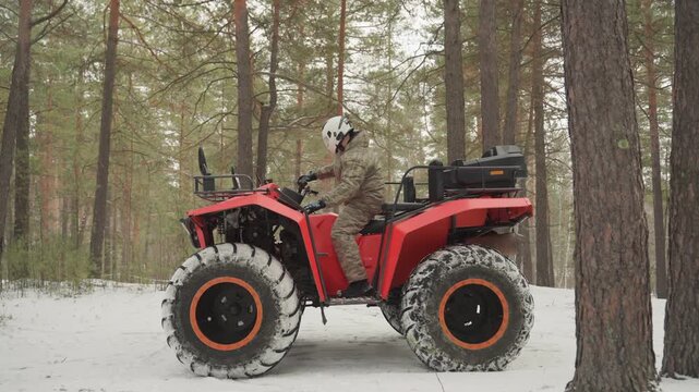 Red atv in snowy forest camo clad rider inspects quad with oversized studded tires snowcovered pine trunks overcast light closeup profile shots show throttle test traction check slow cruising