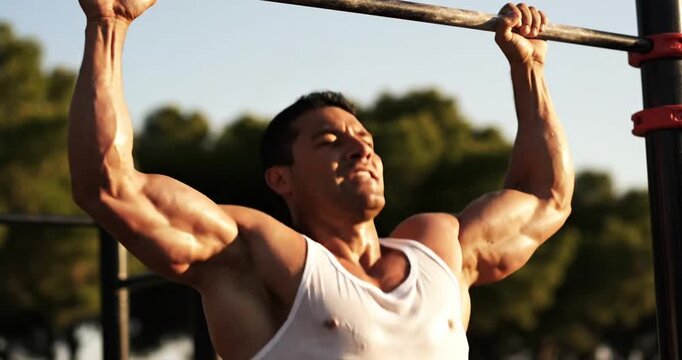 Fit man performing pull-ups in a sunny outdoor gym surrounded by trees and clear sky