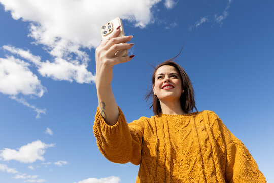 Smiling woman holding smartphone in sunlight over blue sky
