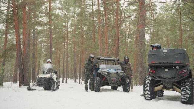 Snowy pine forest atv convoy prepping guide checks gps mechanic inspects engine rider mounts snowmobile beside utv tall pines soft falling snow icy tracks rugged tires teamwork expedition vibe low
