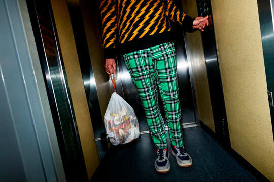 a man in green plaid pants holds a bag of food waste in an elevator
