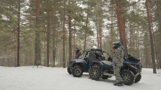 Snow forest atv rider stands beside quad in pine woods, camouflage suit and helmet, soft snowfall, tires on packed snow, waiting to start journey, calm anticipatory mood, adventure prep