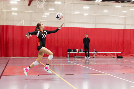 A girl in motion serving a volleyball while her coach watches
