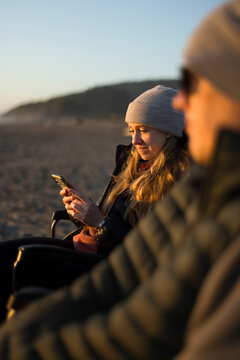A girl and a father sitting on a beach bundled  as she checks phone.