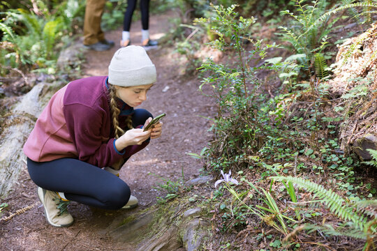 Girl in knit hat photographing a flower on a trail. 