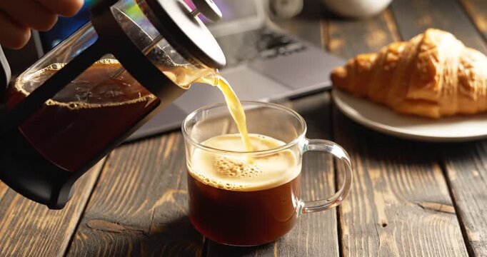Freshly brewed coffee being poured into a glass cup beside a croissant on a wooden table