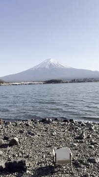Mountain lake scenic view with peaceful fuji captured vertical video frame showing calm water reflecting tranquil atmosphere near shore rocks and serene nature landscape, folding chair stony ground